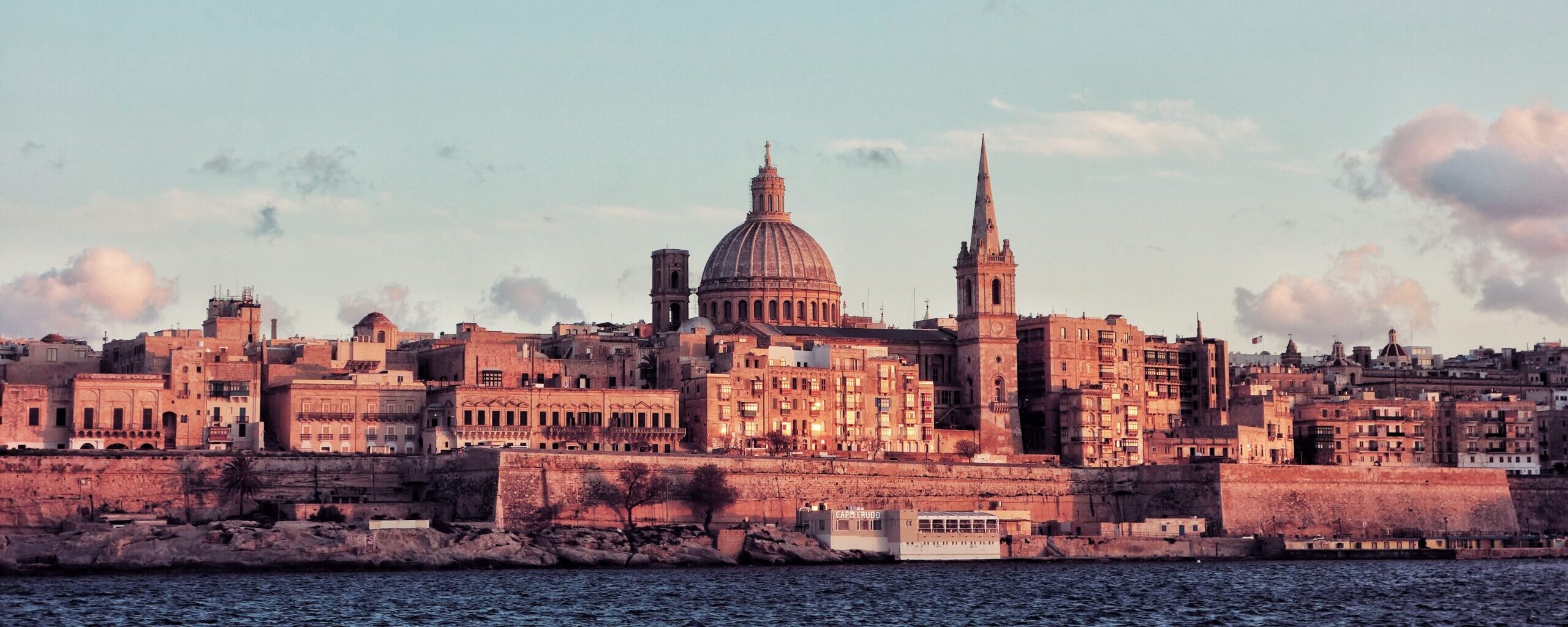 Malta's capital city with the dome and spire of the Carmelite Church and the Basilica of Our Lady of Mount Carmel, along with other historic limestone buildings, overlooking the Grand Harbour.