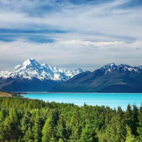 Mount Cook, New Zealand, featuring snow-capped mountains reflecting over a turquoise glacial lake, surrounded by green forests.