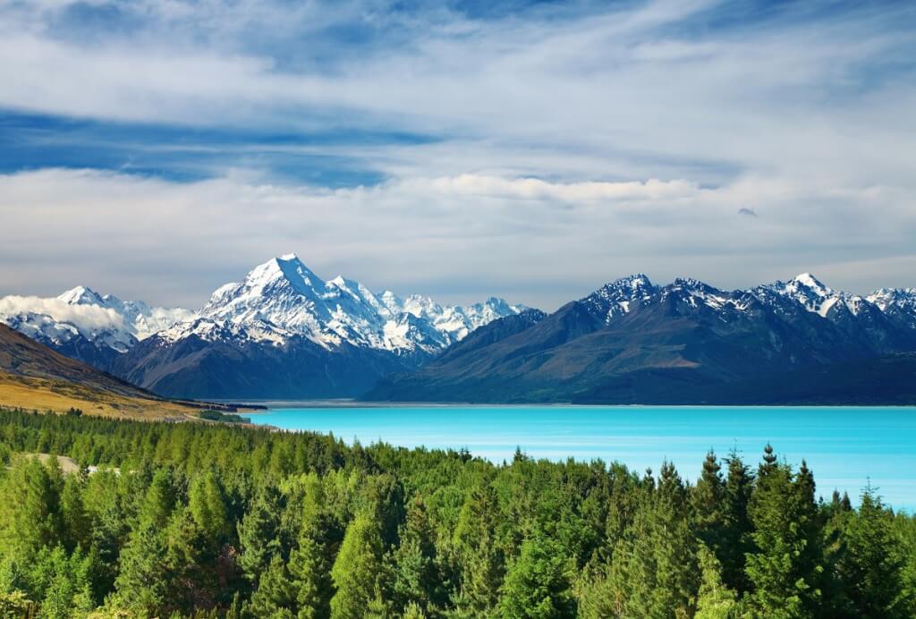 Mount Cook, New Zealand, featuring snow-capped mountains reflecting over a turquoise glacial lake, surrounded by green forests.