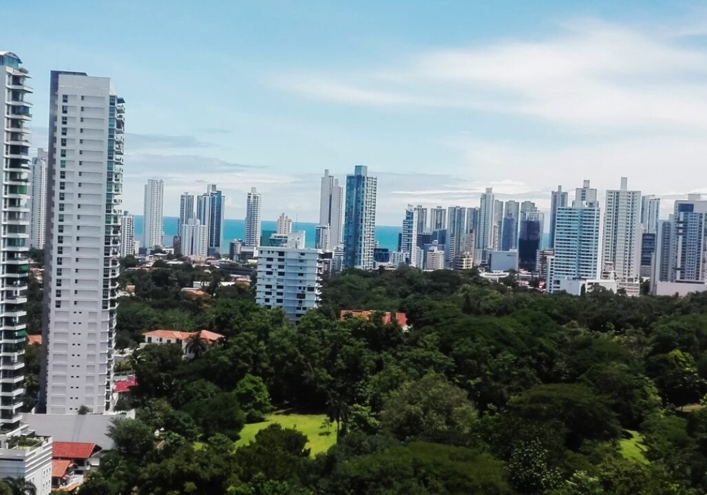 A modern skyline of high-rise buildings in Panama with a green urban park area in the foreground and the ocean visible in the distance.