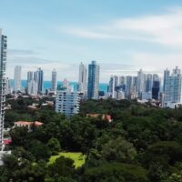 A modern skyline of high-rise buildings in Panama with a green urban park area in the foreground and the ocean visible in the distance.