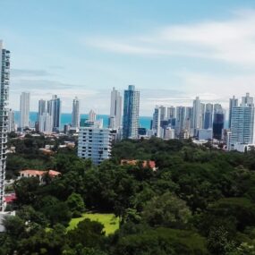 A modern skyline of high-rise buildings in Panama with a green urban park area in the foreground and the ocean visible in the distance.