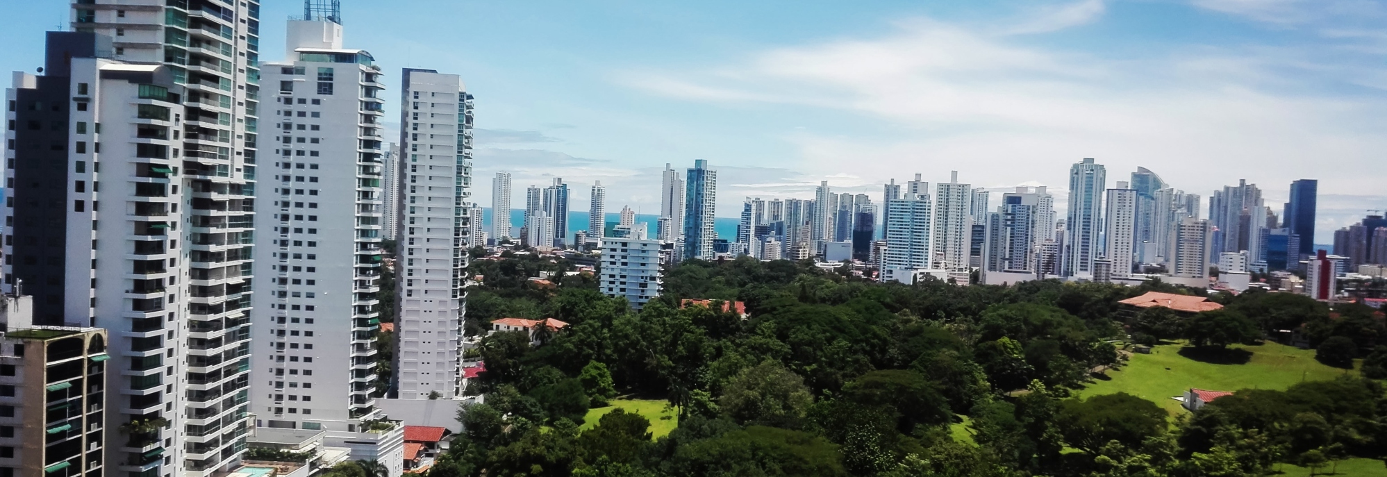 A modern skyline of high-rise buildings in Panama with a green urban park area in the foreground and the ocean visible in the distance.
