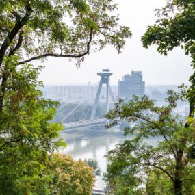 Slovakia, features the UFO Bridge spanning the Danube River, seen through a frame of green trees.