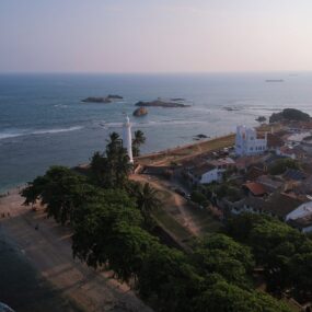 Sri Lanka, a white lighthouse, a fortified wall along the coastline, and historic buildings with red-tiled roofs.