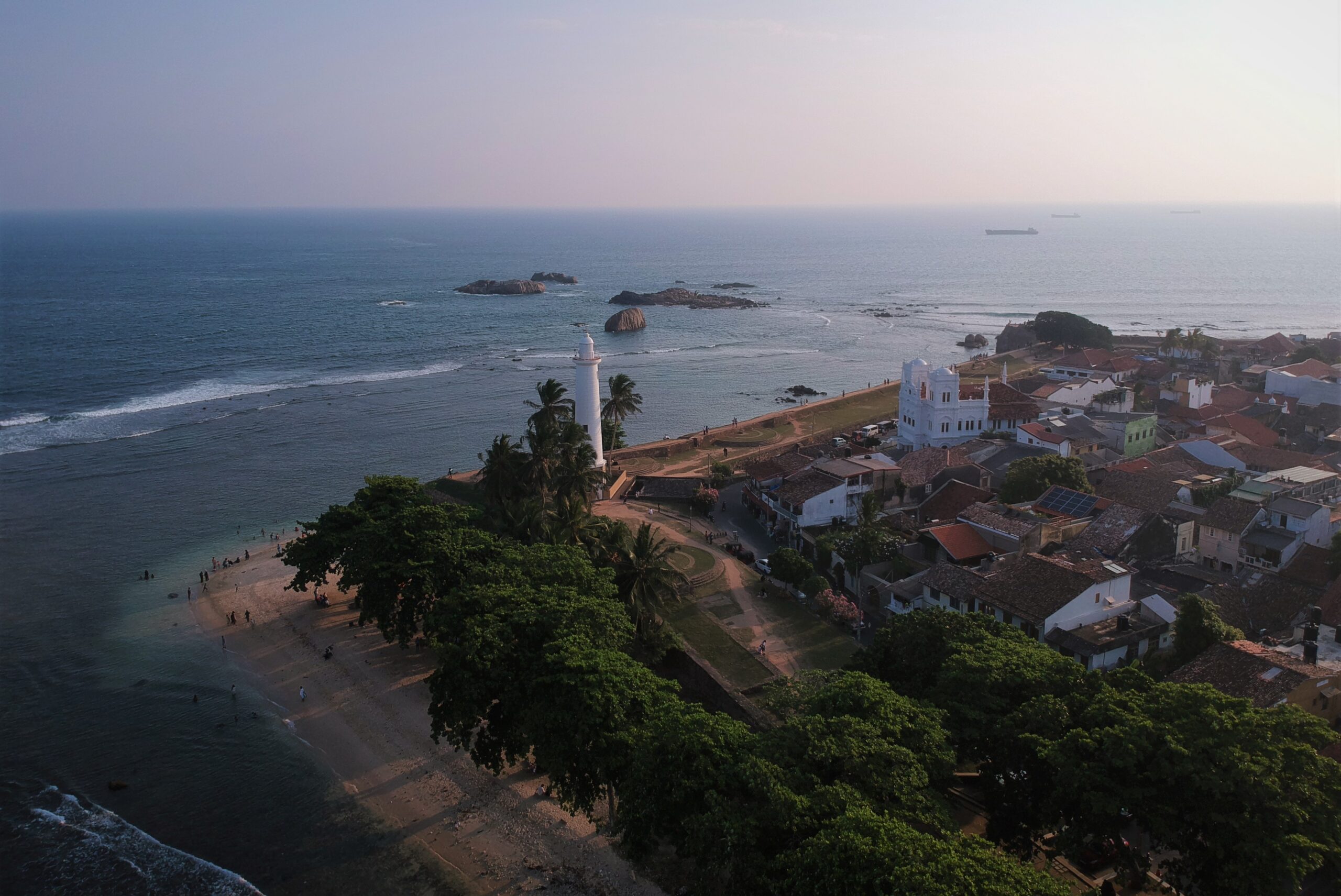 Sri Lanka, a white lighthouse, a fortified wall along the coastline, and historic buildings with red-tiled roofs.