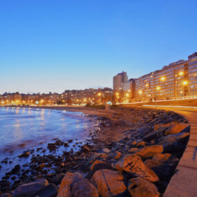 Uruguay, a coastal city at dusk, with a prominent promenade along the water, city lights illuminated, and residential buildings lining the shore.