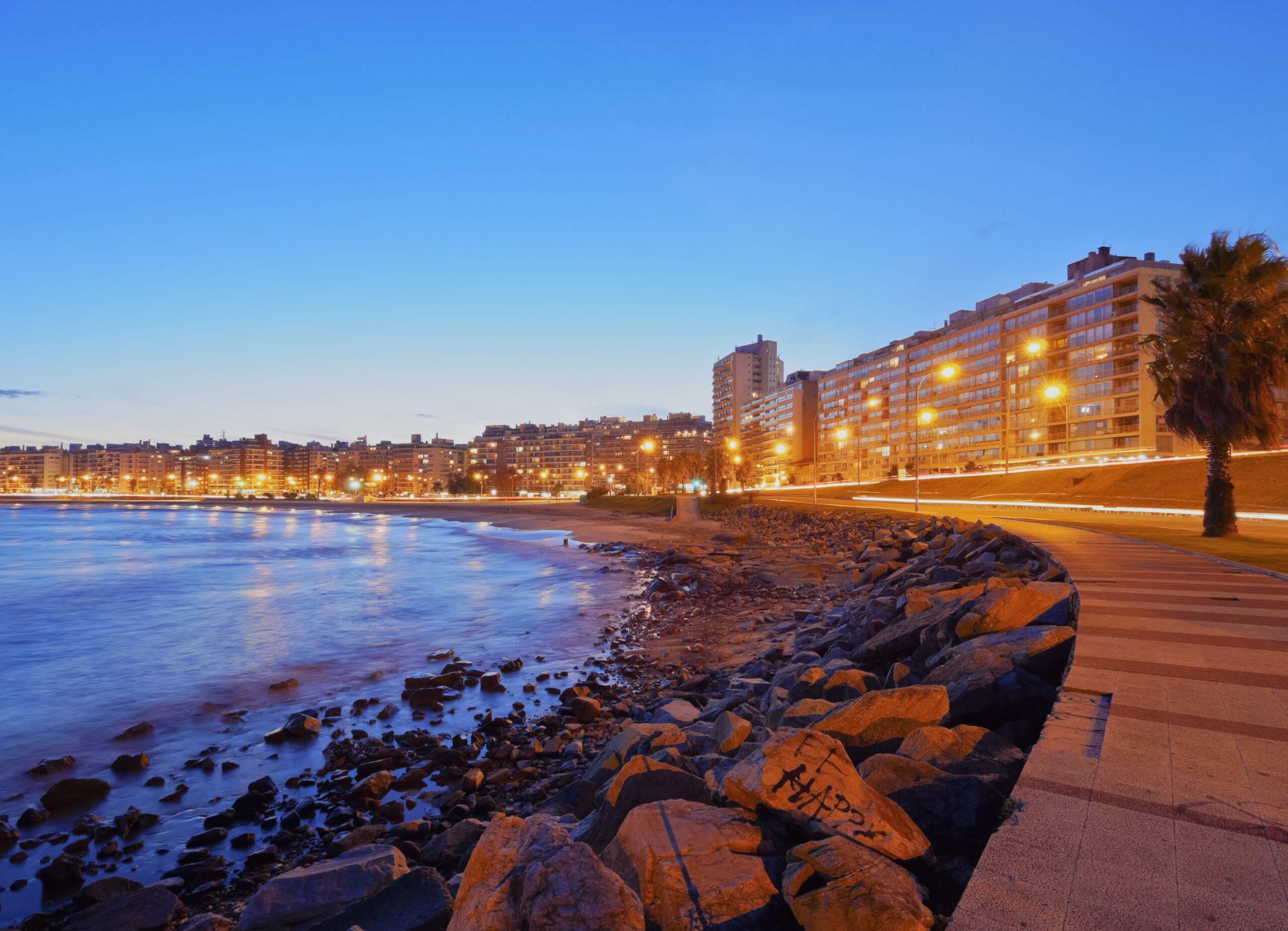 Uruguay, a coastal city at dusk, with a prominent promenade along the water, city lights illuminated, and residential buildings lining the shore.