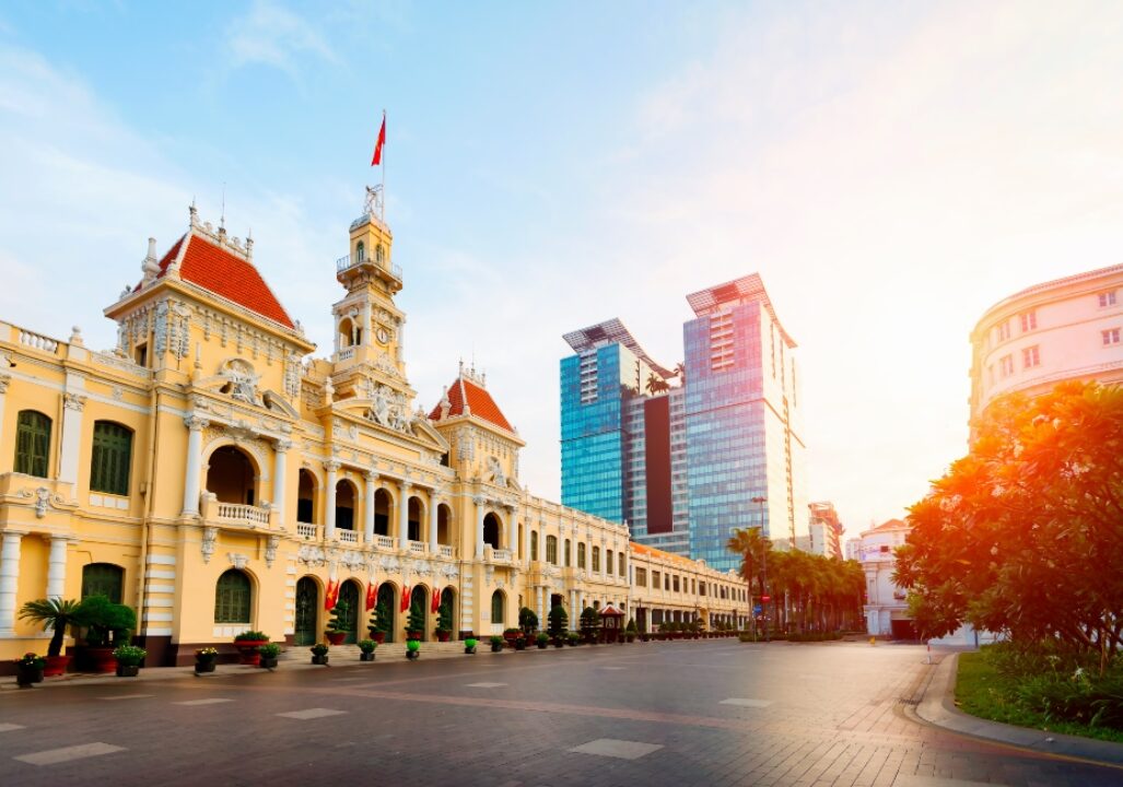 Historic colonial building with red roofs contrasts modern glass skyscrapers under a clear sky. Sunlight casts a warm glow, creating a serene atmosphere.