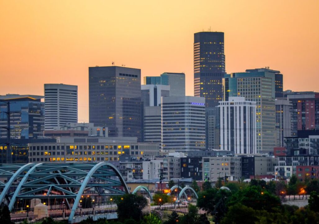 Denver skyline at sunset with tall buildings against a warm orange sky. A blue arched bridge and greenery are visible in the foreground, conveying a calm urban scene.