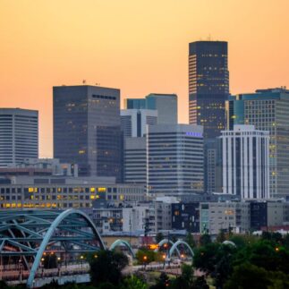 Denver skyline at sunset with tall buildings against a warm orange sky. A blue arched bridge and greenery are visible in the foreground, conveying a calm urban scene.
